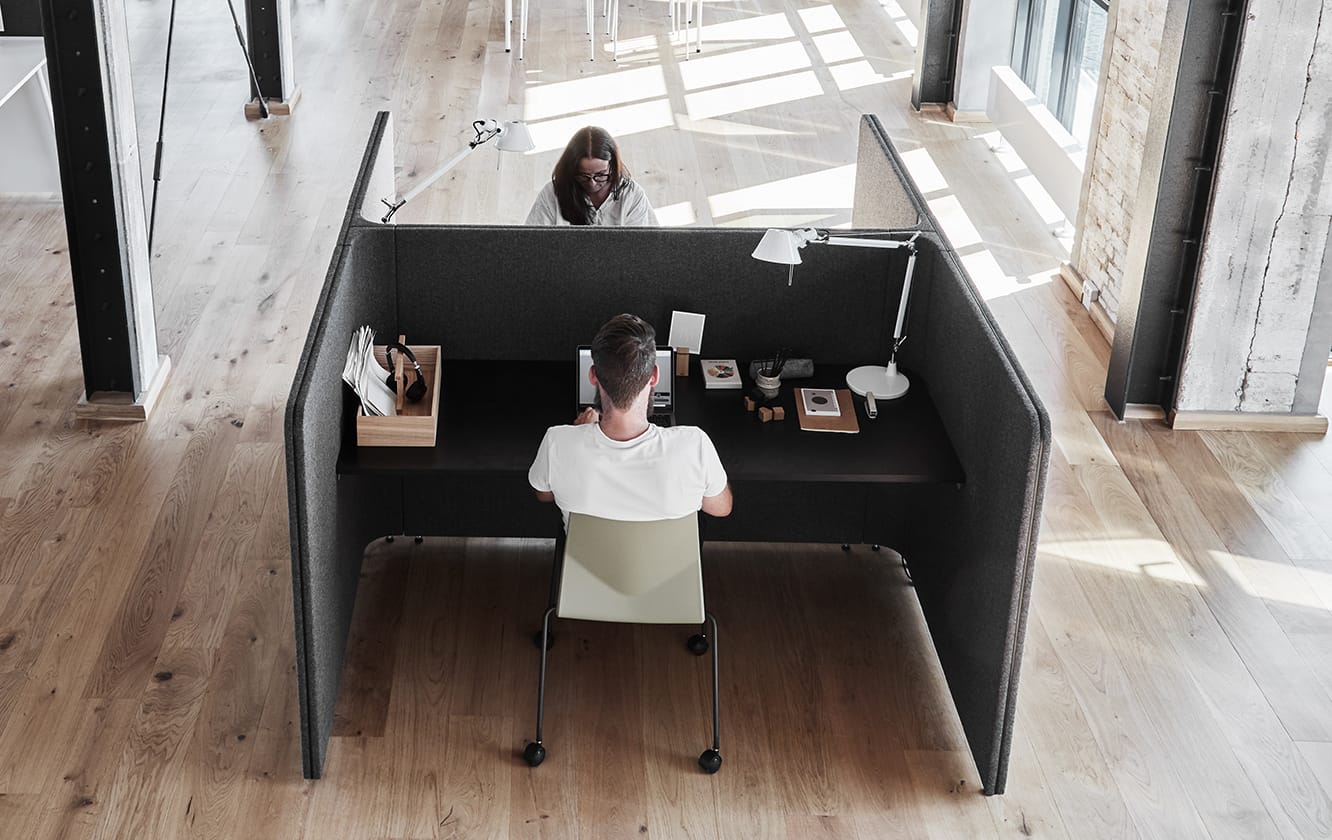 A woman and man sit at a desk in a office work booth with two compartments in an office.