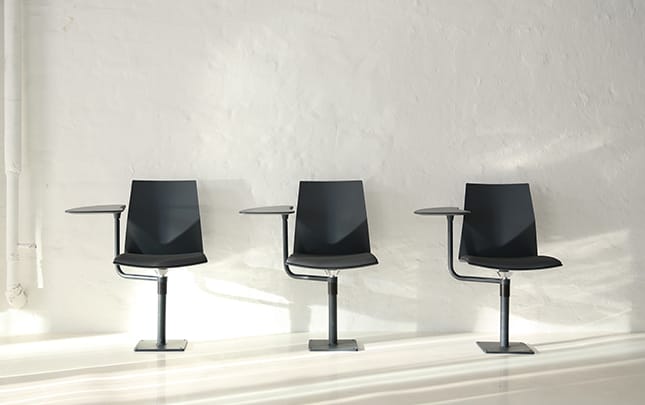 Four black chairs with desks attached in front of a white wall.