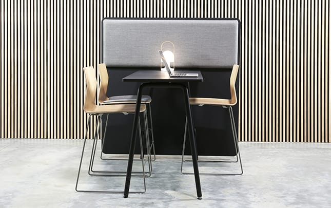 A black table against a office screen divider with an acoustic panel on it and chairs in a room with a striped wall.