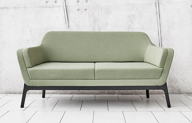 A green couch with black legs in front of a concrete wall.