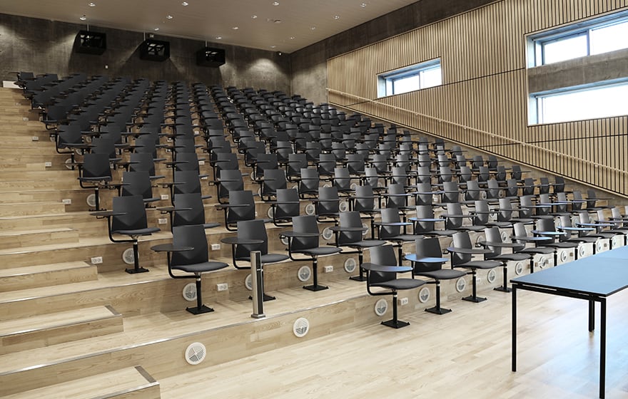 A large auditorium with rows of black chairs with desk attached.