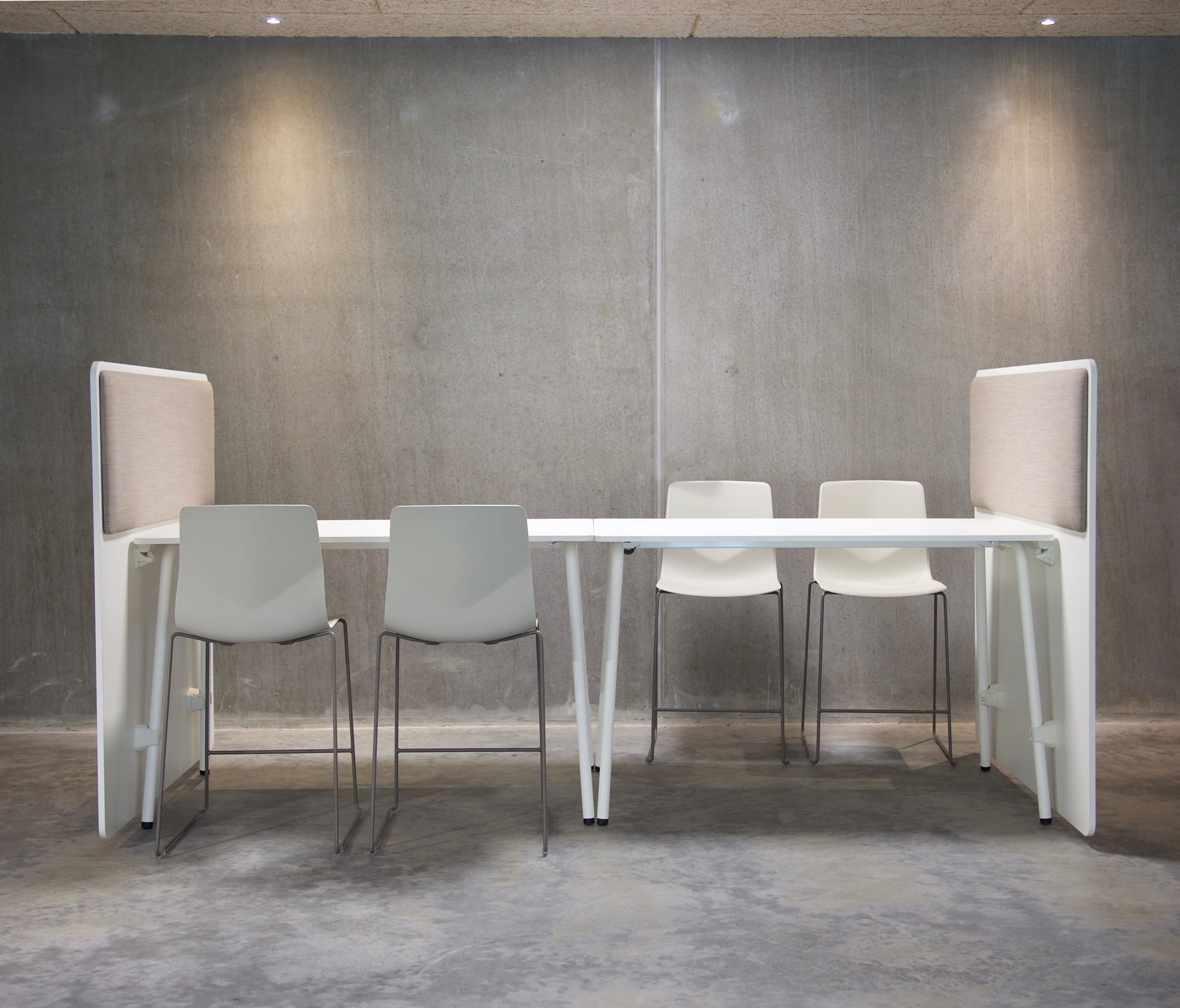 White office tables and counter height chairs seperated by office screen dividers in a room with concrete walls.