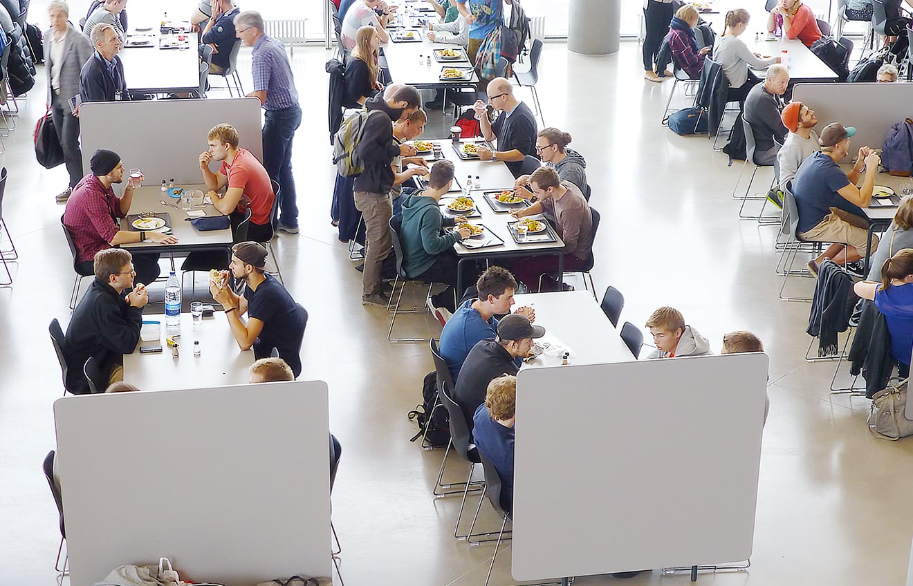 A group of people eating at tables in a large room with office screen dividers partitianing the space