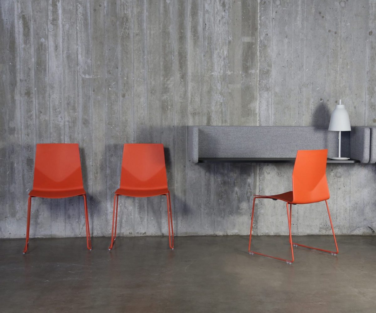 Three orange chairs in front of a desk workstation and concrete wall.
