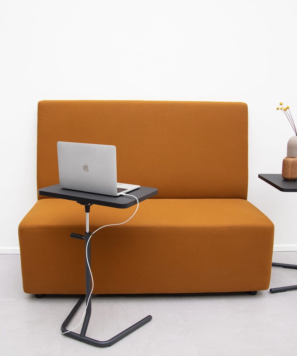 Adjustable height work table with laptop on it in front of an orange office sofa